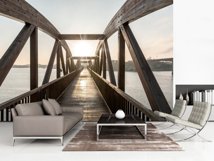 Carta da parati Ponte sul fiume - paesaggio architettonico urbano con un ponte di legno sul fiume con il sole sullo sfondo